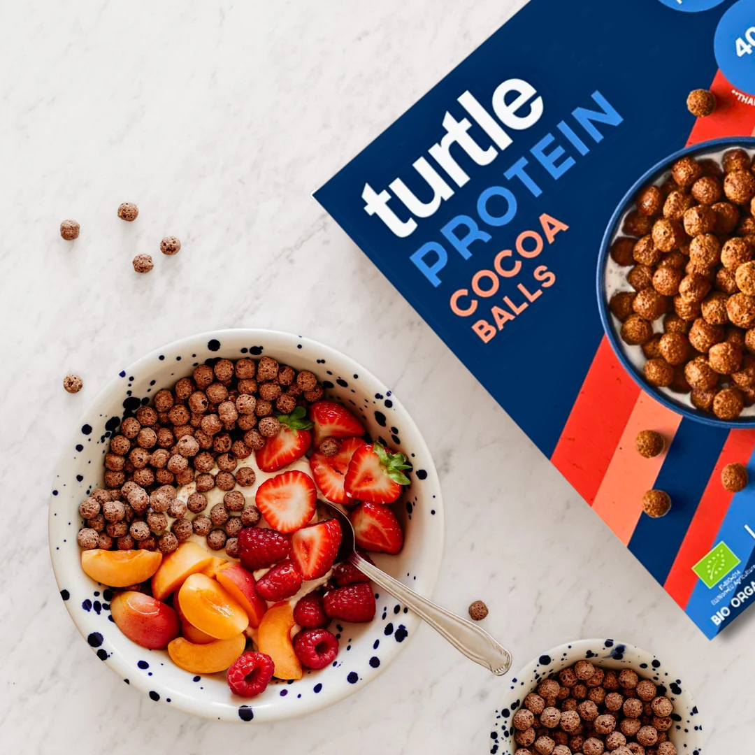 Breakfast bowl with Turtle Protein Cocoa Balls, strawberries, raspberries and apricots next to the cereal box.